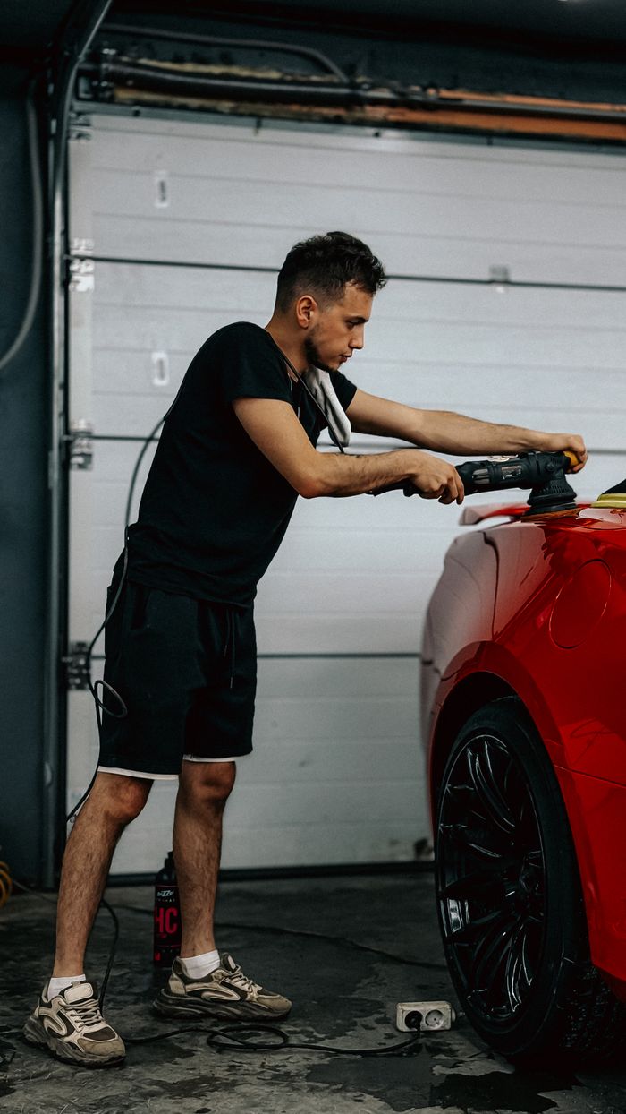 faq-img A man uses a polishing machine on a red car in a garage setting, focusing on detailing.